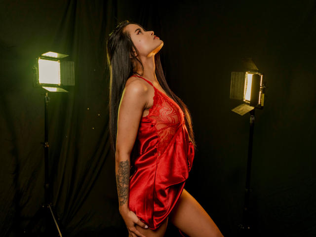 KarolPalacios in a red satin lingerie dress, standing with head tilted back, long black hair, tattoo on right arm, lit by two bright studio lights, showing her ass.