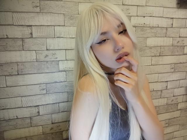 Blonde EileneSzala with long hair, eyeliner, and lip gloss, biting her finger, wearing a gray top, against a textured brick wall. She has a cute, sexy look.