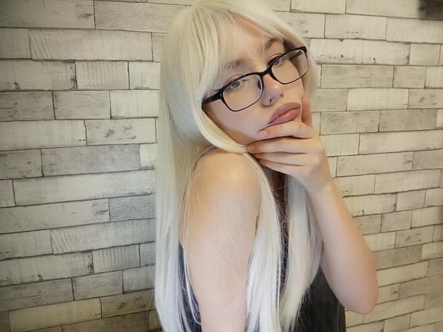EileneSzala with long blonde hair, glasses, and pouty lips, touching her chin, wearing a black top, showing her pale skin and small tits against a wooden wall.