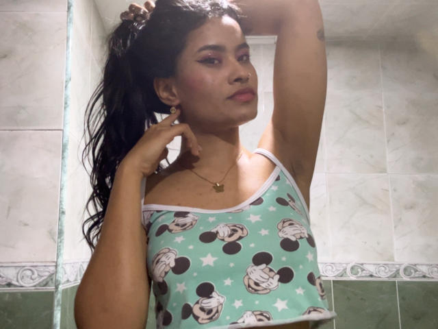 KarolPalacios, con cabello oscuro recogido en una coleta, vistiendo una camiseta verde de Mickey Mouse, con los brazos levantados, mostrando sus pechos, y un collar de oro, luciendo segura de sí misma..