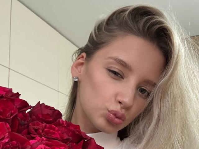 RoseComstock has blonde hair, light skin, and pouty lips, wearing diamond earrings, holding red roses, with a white top, in a selfie.