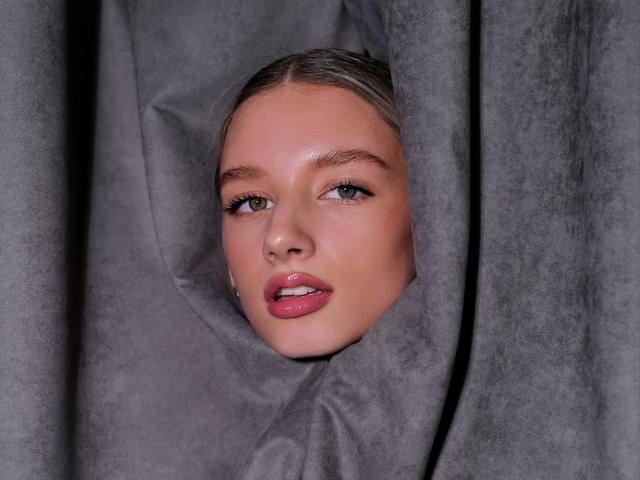 RoseComstock's face peeking through gray curtains, with shiny pink lips slightly parted, showing white teeth, and her green eyes looking forward. Her blonde hair is tucked behind.