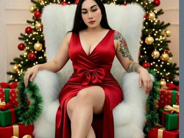 TSweetieCumForU is a curvy woman with large tits in a red satin dress, sitting on a white fur chair, showing a thigh, with a Christmas tattoo on her arm.