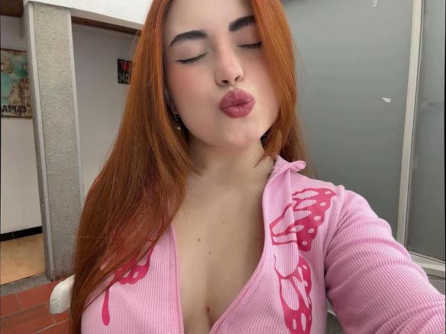 VicluX with red hair, pouty pink lips, and eyes closed, wearing a pink ribbed zip-up with mushroom designs, showing her large tits. She's in a selfie.