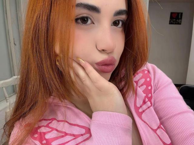 VicluX heeft oranje haar, roze lippen en ze trekt een zuur gezicht met haar hand op haar wang. Ze draagt een roze hoodie met roze patronen. Haar borsten zijn zichtbaar..