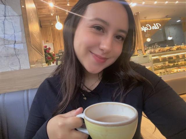 RosalieDupont with dark hair, smiling, holding a yellow and white mug, wearing a black long-sleeve shirt, in a cozy cafe. Her tits are slightly visible.