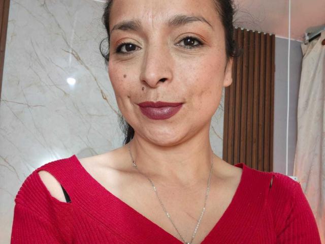 StangelarRivera, een vrouw met een medium huidskleur en donker haar, met kleine moedervlekken op haar neus, draagt een rode trui met uitgesneden schouders en donkerrode lippenstift, en glimlacht naar de camera..