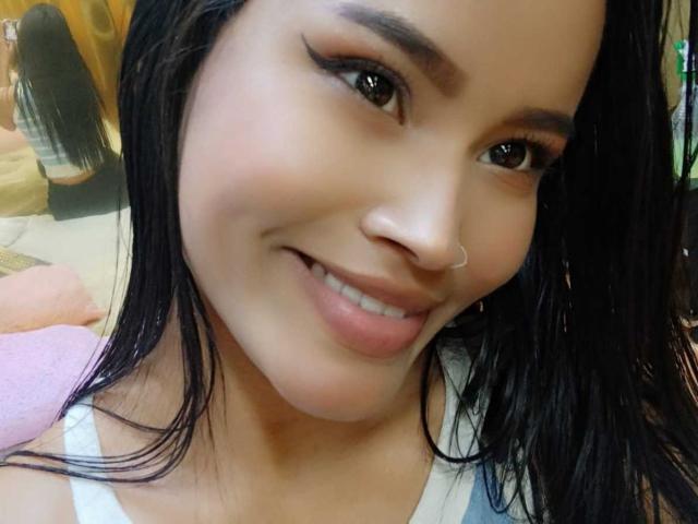 KarolPalacios has medium brown skin, black straight hair, and a small nose ring. She's smiling with full lips, wearing eyeliner, and a white tank top. Her tits are not visible.