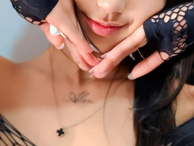 YayaLm's face with pink lips, black lace fingerless gloves, and manicured nails, touching her neck. She has a small tattoo and wears a black cross necklace.