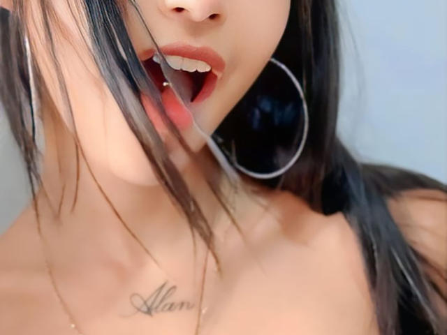 YayaLm with black hair, red lips, and tongue out, wearing a large hoop earring, showing a tattoo below her tits, and a necklace. Her ass is partially visible.