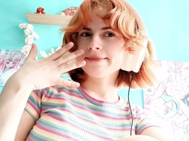 Mireille Spotwood, avec des cheveux courts et orange, portant une chemise rayée, des écouteurs beige, souriante, la main près du visage, un mur bleu vif, des affiches d'anime et des fleurs blanches en arrière-plan..