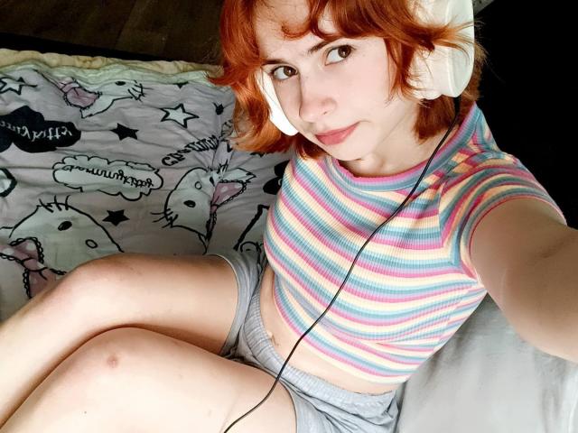 Mireille Spotwood, con cabello rojo, vestida con un top corto a rayas y pantalones cortos grises, sentada en una cama con auriculares, mostrando una cara linda y senos pequeños, con las piernas cruzadas..