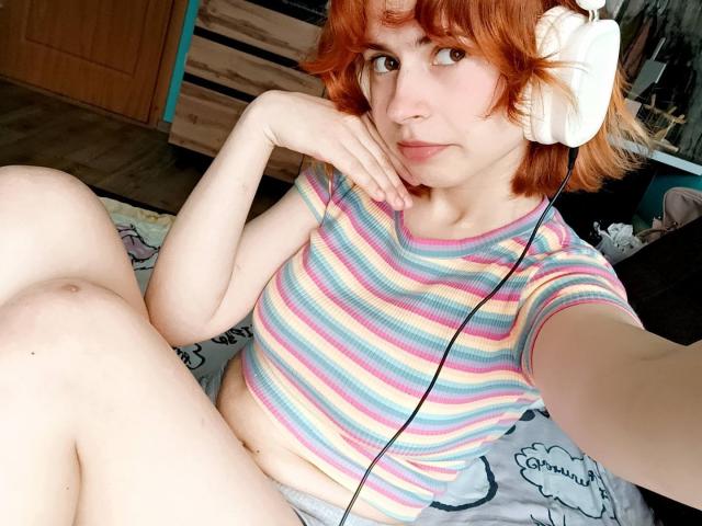 Mireille Spotwood, com cabelo ruivo, usando fones de ouvido brancos, camisa listrada em rosa, azul, amarelo e verde, shorts cinzas, sentada na cama, com a mão no rosto, sorrindo levemente, pernas levantadas..