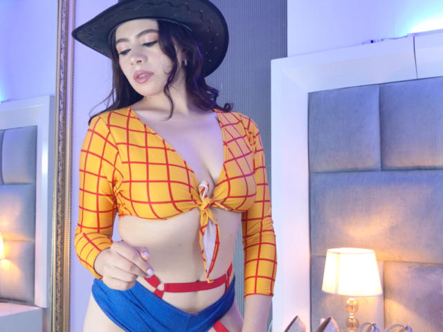 Linda Mancini, con i capelli scuri e ondulati, indossa un cappello da cowboy nero, una maglietta a quadretti gialli e rossi, allacciata per mostrare il seno, e una gonna blu, tirando giù le bretelle rosse..