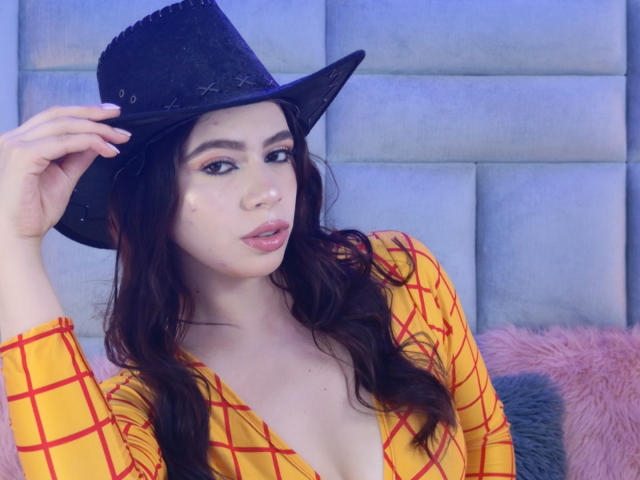 Linda Mancini, con capelli castani ondulati, indossa un cappello da cowboy nero e una camicetta gialla a quadretti, con un taglio basso e un trucco leggero, mentre tocca il suo cappello..