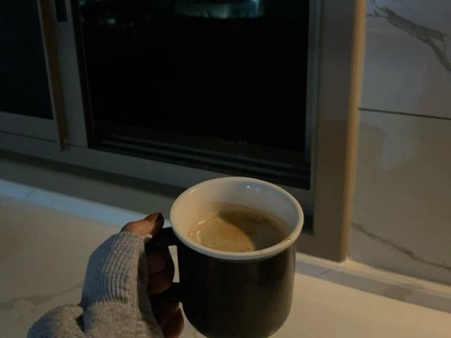 Mindys Hand, die einen grauen Pullover trägt, hält eine schwarze Espressotasse mit dunkler Flüssigkeit im Inneren auf einem weißen Marmelfensterbrett in der Nacht..