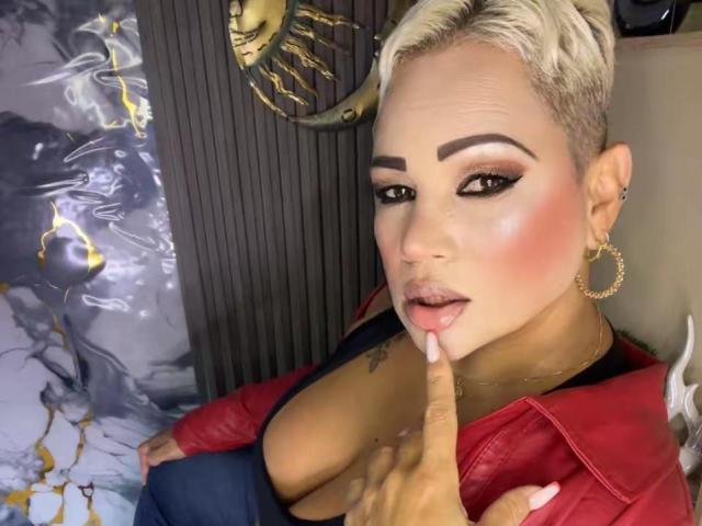 GianellaCox with short blonde hair, red shirt, and blue jeans, finger in mouth, showing off her big tits, pink blush, and gold hoop earrings, with a sexy, confident look.