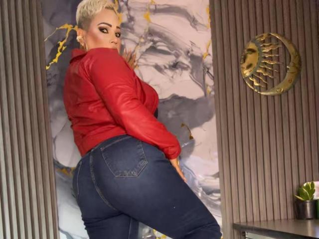 GianellaCox with blonde hair, red shirt, and tight blue jeans, showing her big ass while looking back, standing in a room with a gold wall decoration.