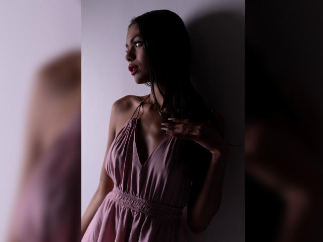 CrystalDavids mit dunklem Haar, trägt ein pinkes Kleid mit tiefem Ausschnitt, steht an einer Wand, Hand auf der Brust, im Schatten, blickt zur Seite, rote Lippen, weiches Licht..