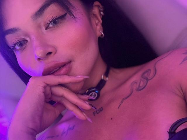 MajoJones with dark hair, purple lighting, eyeliner, long nails, finger near mouth, tattoos on chest, collar with symbol, earrings, slight smile, looking to the side, soft pink glow.