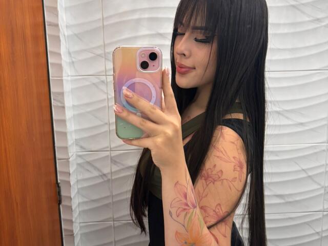 AlisonTatcher with long black hair, wearing a green top, taking a selfie with pink and green phone case, showing tattooed arm with pink and orange flowers.