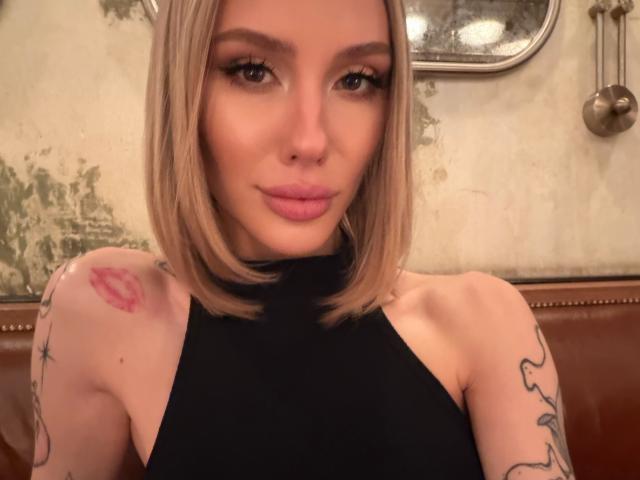 SensualMila with blonde bob, pink lips, black sleeveless top, tattoos on arms, red kiss mark tattoo on shoulder, brown leather bench, looking at camera with subtle smile.
