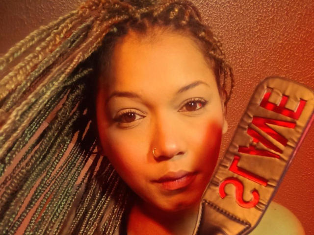 EmperatrizFetishist a la peau brune, de longs cheveux tressés, un anneau au nez et un léger sourire, et elle tient un panneau rouge avec le mot "HAPPY" écrit en lettres grasses..