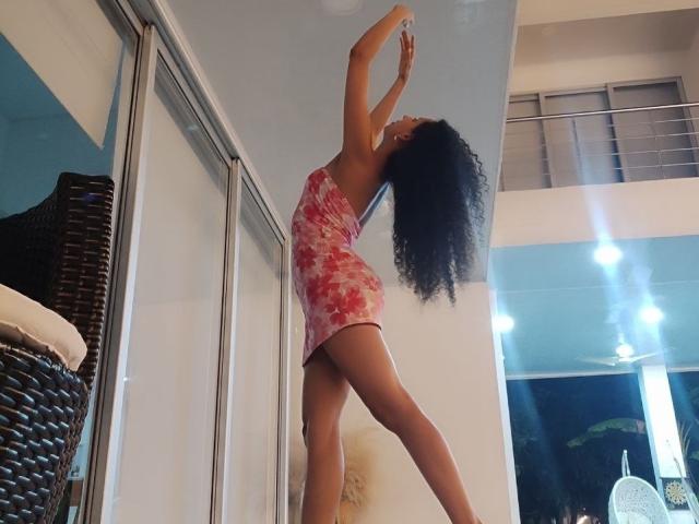 CurlyBlossom with curly black hair, wearing a pink floral dress, stretching arms upward, showing her ass and legs, standing in a bright room with mirrors and wicker furniture.