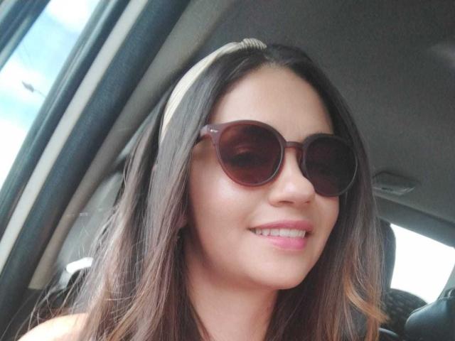 TheresaLarson with brown, oval sunglasses, white headband, and pink lipstick, smiling slightly, showing straight white teeth, in a car.