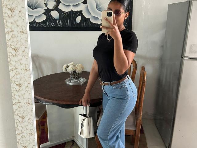 NatalyBlonder, avec de grandes seins, portant un t-shirt noir, un jean bleu taille haute, tenant un sac blanc, debout devant un miroir, en train de prendre un selfie..
