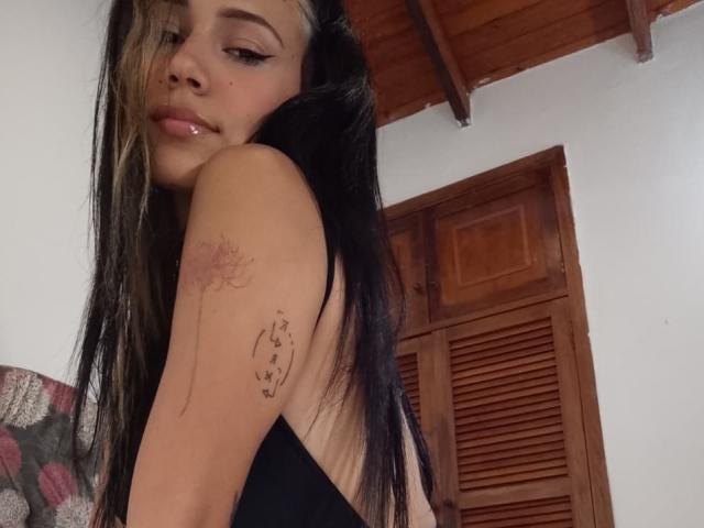 KataleyaMansson with long black and blonde hair, glossy pink lips, black tank top, tattoo on right arm, looking over shoulder, wooden ceiling and shutters in background.