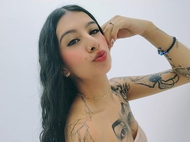 CarlaCorinas, avec ses longs cheveux noirs, son anneau au nez et ses tatouages, lèvres boudeuses, touchant sa joue, portant un collier, un bracelet et une bague en or, montrant ses seins et ses bras tatoués..