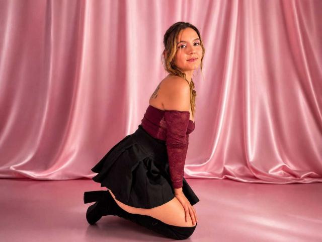 Zoe Collinson kniet auf einem rosa Boden, trägt ein burgunderfarbenes Oberteil mit freier Schulter, einen schwarzen Rock und schwarze High Heels. Dabei zeigt sie ihr Gesäß und ein kleines Tattoo an ihrer rechten Schulter..