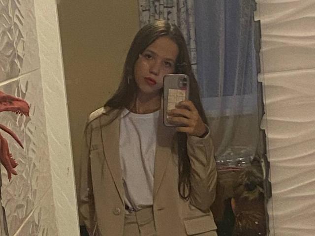 KarenPortal, avec de longs cheveux bruns, portant un costume beige et une chemise blanche, tenant un téléphone, autoportrait dans un miroir, rouge à lèvres, main droite levée, expression subtilement confiante, reflet réaliste dans le miroir..