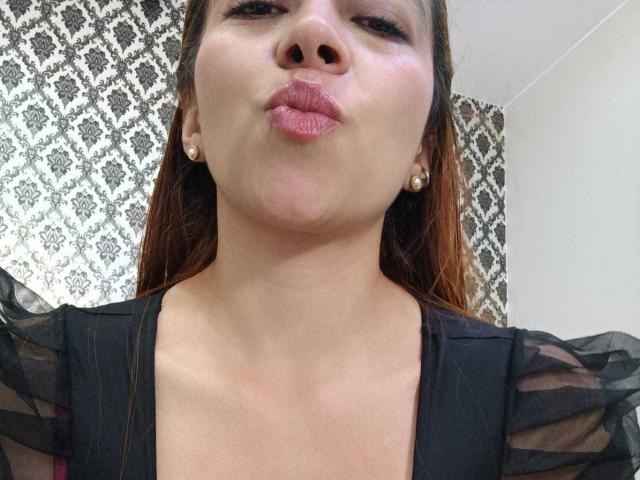 MadisonKoper with puffy pink lips, wearing a black sheer top, small pearl earrings, brown hair, and a slightly upturned nose, making a puckering face.