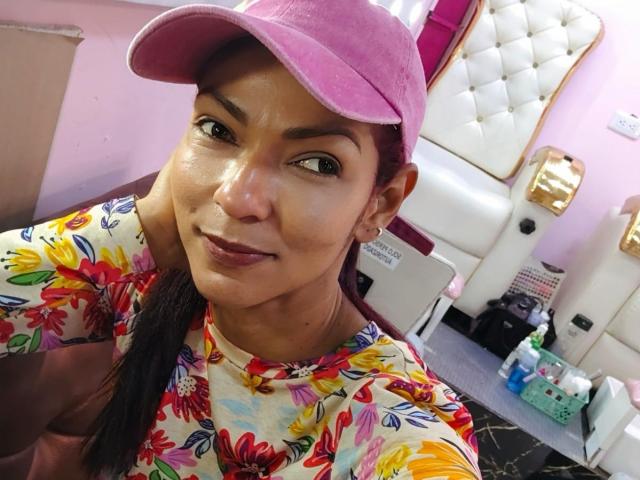 HannaPretty with shiny skin, brown eyes, and pink cap, wearing floral shirt, smiling at camera. Boobs not visible, pussy not visible, ass not visible. Room with white tufted chair.
