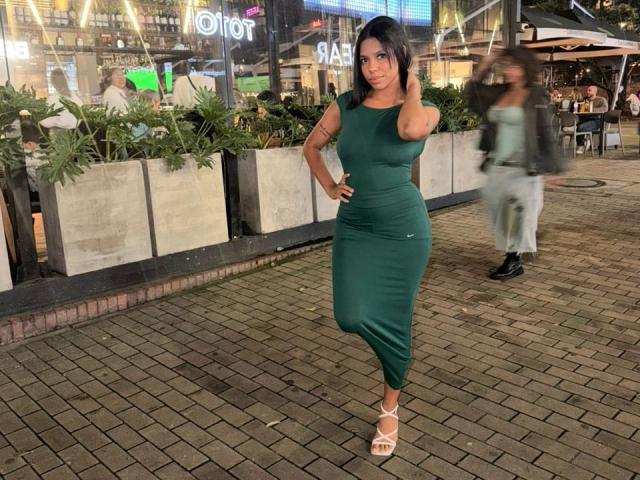SammyRosi, con un vestido verde ajustado, de pie con una mano en la cadera, mostrando su trasero, con tacones blancos de tiras, con su cabello oscuro suelto, en una escena nocturna de la ciudad..