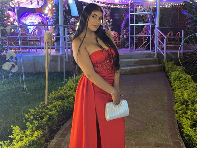 AlaraKey with long black hair, red strapless dress showing her big tits, holding a white clutch, standing on a brick path, night, outdoor, festive lights.