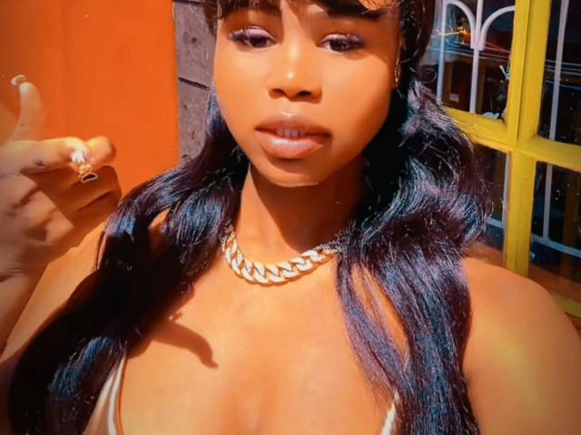 EbonyStar-hot with dark brown skin, glossy black hair, and a silver chain necklace, showing off her shiny lips and small, round tits. She's giving a thumbs-up.