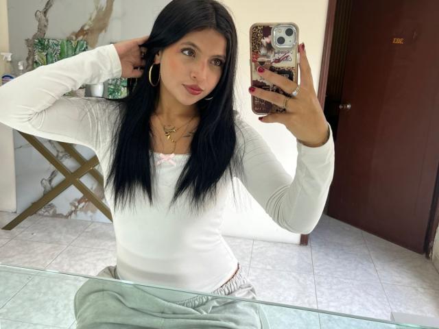 NyraVanel with long black hair, wearing a white long-sleeve top and green pants, taking a mirror selfie with a marble-patterned phone, showing her tits and small ass.