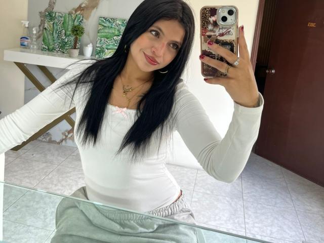 NyraVanel with long black hair, white long-sleeve top, pink bow, gray pants, holding phone, smiling, sitting, mirror selfie, visible tits, pink nails, gold necklace, white tiled floor.