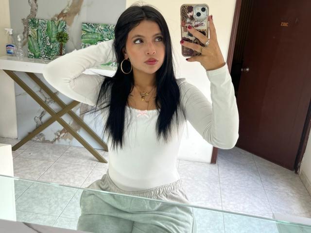 NyraVanel with long black hair, wearing a white long-sleeve top and gray pants, taking a mirror selfie with her right hand in her hair, showing her tits slightly.