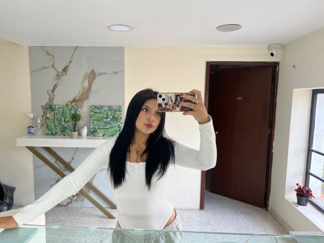 NyraVanel with long black hair, white long-sleeve top, and pink underwear, holding a phone with a patterned case, taking a mirror selfie, showing her small tits and ass.