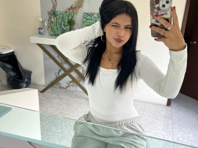 NyraVanel with black hair, white long-sleeve shirt, gray pants, holding phone in mirror selfie, hand in hair, pink bow, red nails, small tits, glass table, white wall.