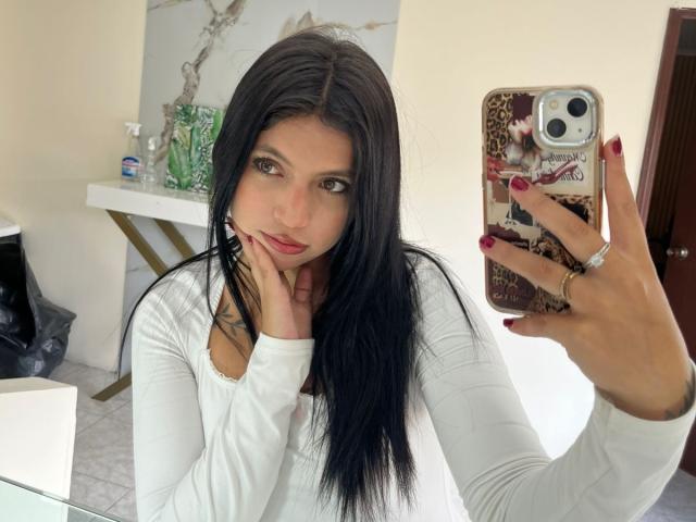 NyraVanel with long black hair, white long-sleeve shirt, red nail polish, holding phone with leopard print case, hand on chin, looking at mirror, ring on finger.