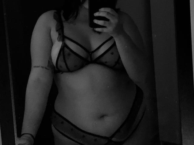 RouseKirryHot en lingerie noire transparente avec des pois, montrant ses seins et son ventre, tenant un téléphone pour prendre un selfie devant un miroir, avec un tatouage sur son bras droit. Photo en noir et blanc..