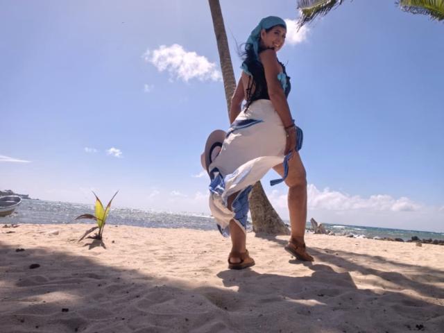 JackieMature stands on a sandy beach, wearing a blue and white headscarf, black top, and white dress, with a blue sash, showing her ass and brown sandals.
