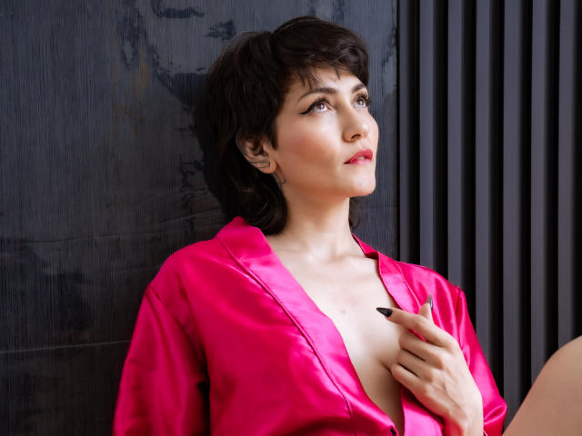 Ivette Durand, met kort donker haar, een roze satijnhemd, een diep decolleté, de rechterhand op de borst, zwarte nagels, die omhoog kijkt, licht geopende lippen, bleke huid, tatoeages op het linker oor..