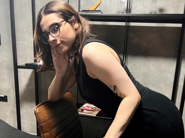 Lia, met een bril, zwarte jurk en bruin bob kapsel, leunt op een bruine stoel, toont een tatoeage op haar rechterarm, met haar hand op haar kin, sexy, schattig, kleine borsten, subtiele make-up, zelfverzekerde glimlach..