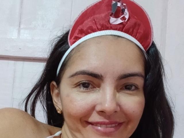 Luaunt with medium brown skin, dark wavy hair, wearing a red satin headband with a white trim and a small red ribbon, smiling at the camera. Boobs not visible.